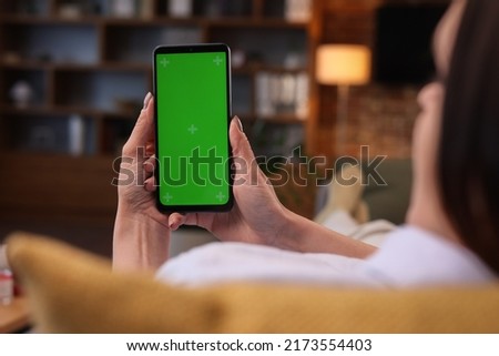 Over shoulder view of young woman holding smartphone with green mock-up screen horizontal mode.Woman lying on couch at home in evening, watching content videos blogs, news or films.Cinematic lighting.