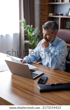 Portrait of adult gray-haired bearded businessman working with laptop at home office. Texting messages, doing paperwork, calculating monthly expenses. Business concept.