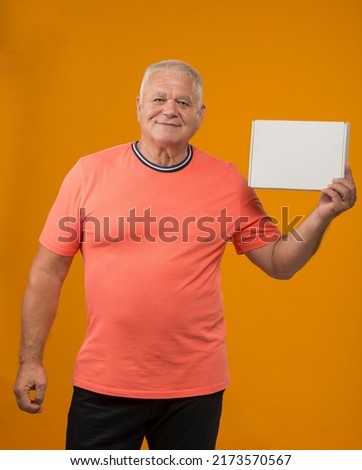 pleasant senior man with a box on a yellow background isolated.an elderly cheerful man in a coral t-shirt shows a box without inscriptions.man holding blank sign