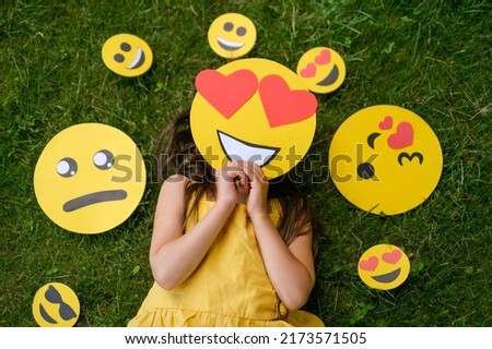 The person is lying on the grass covering his face with a loving emoticon with big hearts instead of eyes. Smile faces with different moods next to the child
