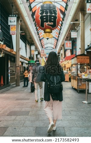 rear view with full length shot asian woman walking under big red lantern hanging overhead along the arcade in kuromon ichiba market in Osaka japan. translation:â kuromonâ Royalty-Free Stock Photo #2173582493