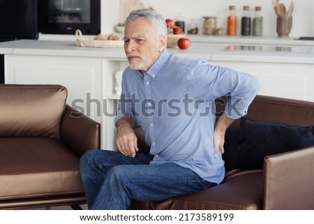 Side view photo of suffering senior man feeling lower back pain, sitting on comfortable orthopedic armchair in incorrect pose. Chronic injury, muscle inflammation and backache concept