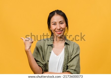 Portrait of young Asian lady smiling with cheerful expression, shows something amazing at blank space in casual cloth and looking at camera isolated over yellow background. Facial expression concept.