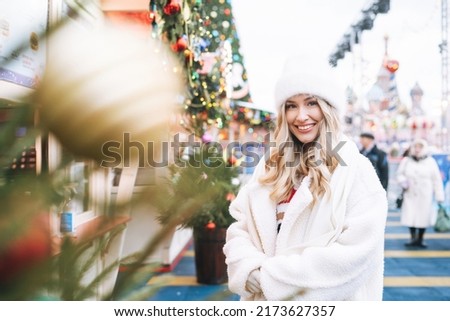 Young happy woman with curly hair in white knitted hat on shopping at Christmas fair market in winter street decorated with lights