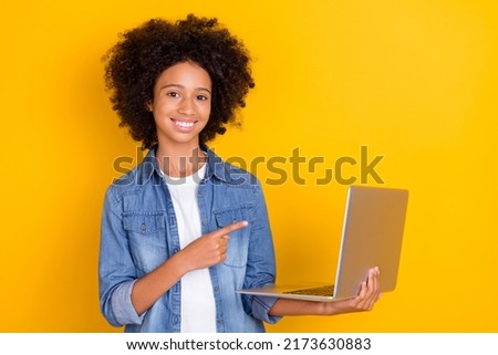 Portrait of beautiful trendy cheerful teen girl holding demonstrating laptop isolated over bright yellow color background