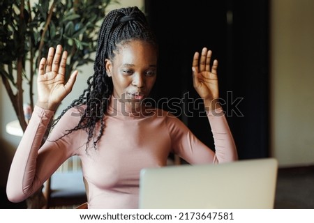 Portrait of young excited African-American woman with long dark braids wearing pink roll-neck, sitting at table in cafe near laptop, raising hands up, expressing success, win. Technology, internet. 