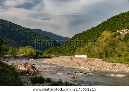 View of the mountain river on a sunny summer day under a blue sky with white clouds. The shallow river flows against the background of the shore, covered with dense vegetation with green foliage.