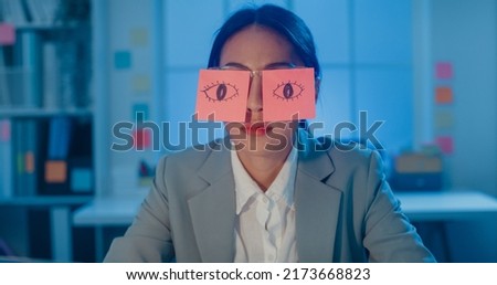 Young Asian businesswoman sitting on desk overworked tired sleep over a laptop at office at night. Exhausted burnout lady with two post-it over her eyes, adhesive notes on face sleeping at workplace. Royalty-Free Stock Photo #2173668823