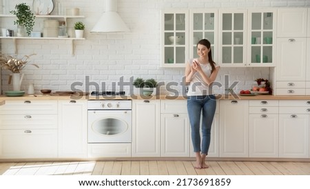 Happy woman surfing net on mobile standing barefoot on warm floor in Scandinavian kitchen. Young housewife in her 20s shopping online using smartphone leaning on modern kitchen countertop and smiling Royalty-Free Stock Photo #2173691859