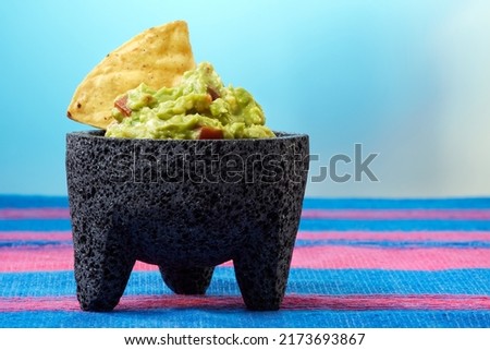 Delicious traditional Mexican avocado guacamole with tomatoes and a nacho or totopo in a typical mortar called molcajete over a blue and pink tablecloth and a blue background. Royalty-Free Stock Photo #2173693867