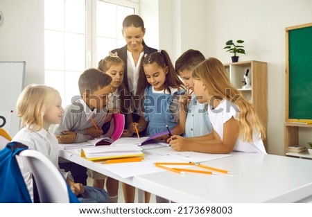 Happy teacher and students having interesting class. Group of school children standing at table in modern classroom, writing with pencils, drawing pictures, checking homework, and learning new skills
