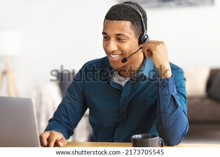 African american young man customer support call center operator or receptionist sitting at the workplace in a modern office consulting a client, uses a headset, smiles friendly Royalty-Free Stock Photo #2173705545
