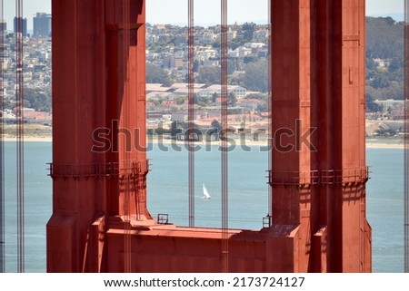 A closeup of one of the red towers of the Golden Gate Bridge (pictured with selective focus) with a sail boat in the background framed by the bridge's support cables in San Francisco, California, USA.