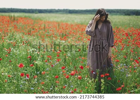 Cottagecore aesthetics. Stylish woman in rustic dress walking in red poppy field. Young female in linen dress relaxing among wildflowers meadow in sunny summer countryside, rural slow life