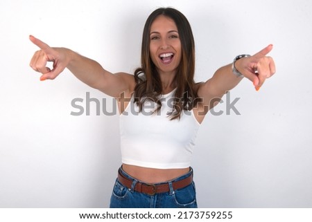 Close-up portrait of surprised young beautiful caucasian woman wearing white top over white background pointing with two fingers to the camera saying: I choose you!, looking up with open mouth.
