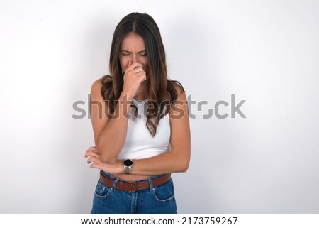 young beautiful caucasian woman wearing white top over white background, holding his nose because of a bad smell.