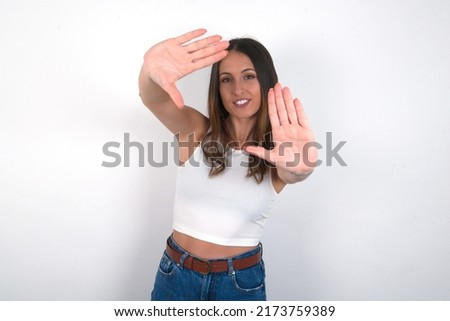 Portrait of smiling young beautiful caucasian woman wearing white top over white background looking at camera and gesturing finger frame. Creativity and photography concept.
