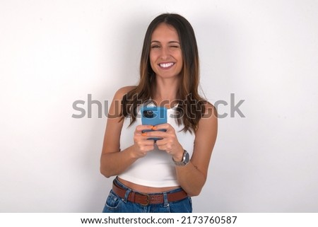 Pleased young beautiful caucasian woman wearing white top over white background using self phone and looking and winking at the camera. Flirt and coquettish concept.