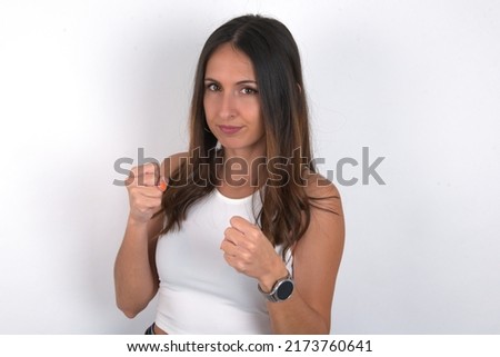 Portrait of attractive young beautiful caucasian woman wearing white top over white background holding hands in front of him in boxing position going to fight.