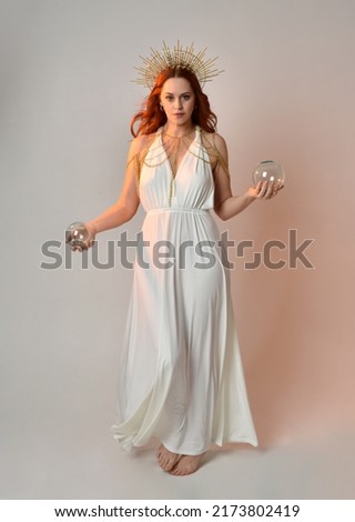 Full length portrait of beautiful red head woman wearing long flowing fantasy toga gown with golden halo crown jewellery, standing pose   isolated on a white studio background.