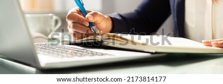 Close-up Of A Businesswoman's Hand Writing Note With Pen In Diary Over Desk