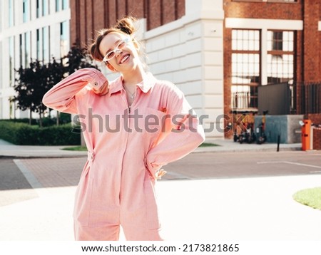 Young beautiful smiling hipster woman in trendy summer pink overall. Carefree teen model posing in the street background at sunset. Positive female outdoors in sunglasses