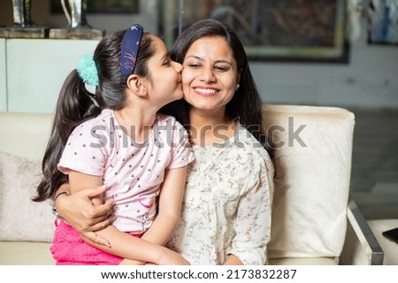 Happy little indian daughter kiss her young mother on cheeks at home.