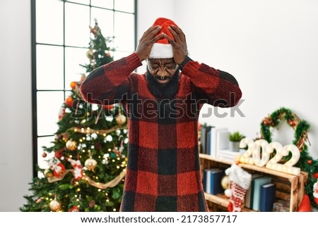 African american man wearing santa claus hat standing by christmas tree suffering from headache desperate and stressed because pain and migraine. hands on head. 
