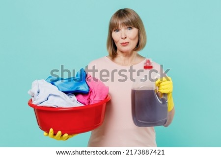Elderly housewife woman in pink t-shirt gloves give stain remover cleanser hold clothes in basin after washing isolated on plain pastel light blue background. Housekeeping cleaning tidying up concept