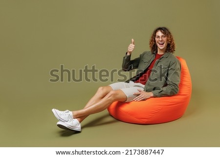 Full size body length fun jubilant young brunet curly man 20s wears khaki shirt jacket glasses sit in bag chair showing thumb up like gesture isolated on plain olive green background studio portrait Royalty-Free Stock Photo #2173887447