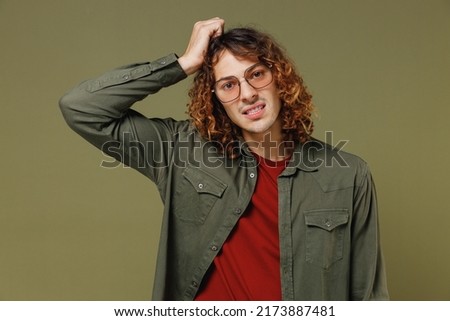 Confused disconcerted unnerved aggrieved upset young brunet curly man 20s wears khaki shirt glasses looking camera put hands on head scratching isolated on plain olive green background studio portrait Royalty-Free Stock Photo #2173887481
