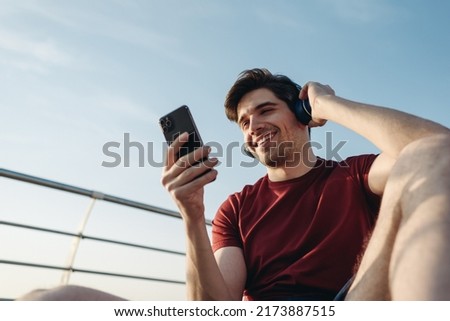 Young smiling strong sporty fit sportsman man in sports clothes headphones sit on pier use mobile cell phone warm up train at sunrise sun dawn over sea beach outdoor on seaside in summer day morning Royalty-Free Stock Photo #2173887515