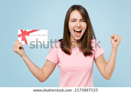 Young excited happy caucasian woman 20s in pink t-shirt hold gift certificate coupon voucher card for store do winner gesture isolated on pastel plain light blue background. People lifestyle concept