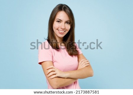 Young smiling cheerful happy fun caucasian woman 20s wearing pink t-shirt look camera hold hands crossed folded isolated on pastel plain light blue background studio portrait. People lifestyle concept
