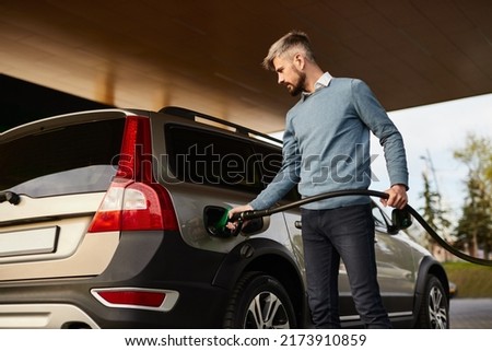 Low angle of concentrated young bearded male driver in smart casual clothes filling fuel into modern SUV car with gas pump nozzle at station Royalty-Free Stock Photo #2173910859