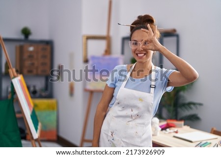 Brunette woman painting at art studio smiling happy doing ok sign with hand on eye looking through fingers 