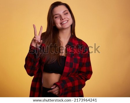 Beautiful lady hold arm hand fingers show v-sign say hi friends  wear casual checkered plaid shirt clothes outfit isolated yellow background. People sincere emotions lifestyle concept.
