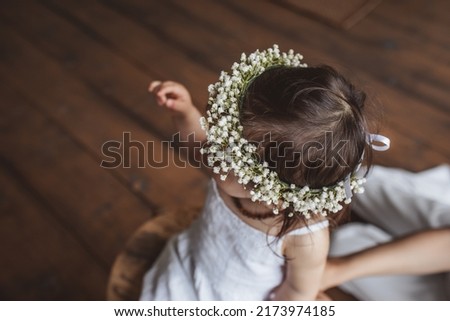 Portrait of pretty one year girl in white summer dress, flower crown and with eco beads sitting in the studio near mom's legs. Eco style concept Royalty-Free Stock Photo #2173974185