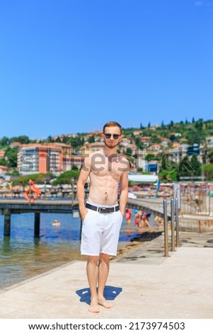 young bearded guy in sunglasses and hold hands in pockets. guy with strong torso stands on the pier on the background of the city