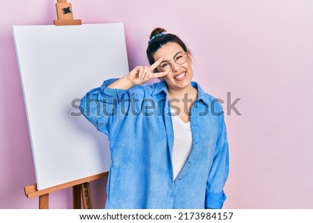 Young hispanic woman standing by painter easel stand doing peace symbol with fingers over face, smiling cheerful showing victory 
