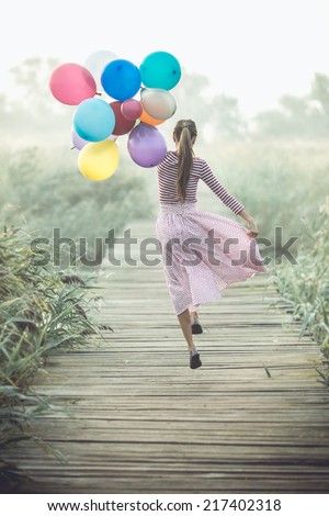 Beautiful woman with colorful balloons running on wooden bridge road early in foggy morning. toned dramatic image, grain added