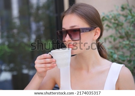 portrait of beautiful attractive happy positive smiling girl in sunglasses, young woman sitting outdoors on terrace in cafe on table enjoying drinking beverage tea coffee at summer sunny day 