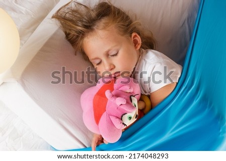A cute blonde Caucasian girl sleeping with a toy on a mattress under a blue cozy blanket