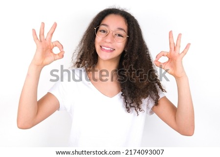 Glad Young beautiful girl with afro hairstyle wearing white t-shirt over white wall  shows ok sign with both hands as expresses approval, has cheerful expression, being optimistic.