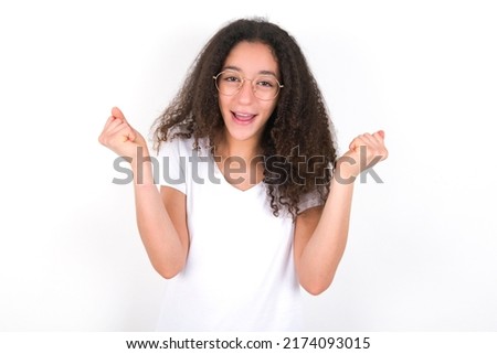 Young beautiful girl with afro hairstyle wearing white t-shirt over white wall  rejoicing his success and victory clenching fists with joy being happy to achieve aim and goals. Positive emotions, feel