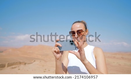 Tourist woman using cell phone for shooting, standing in desert. Action. Young female taking picture of desert on her Iphone.
