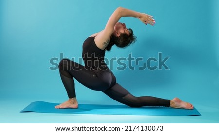 Fit adult recovering from workout practice with stretching exercise in studio. Athletic woman doing body stretch activity after pilates training with fitness equipment over blue background. Royalty-Free Stock Photo #2174130073