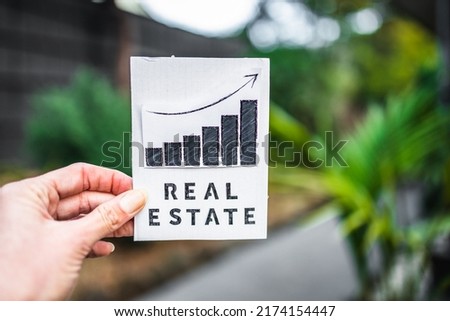 hand holding real estate sign with graph showing stats going up in front of backyard bokeh, concept of property value and return on investment or rent price