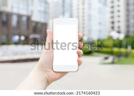 phone mockup with blank white screen. A female hand holds a white smartphone with a blank screen on a background of buildings. Woman showing empty phone screen. Mockup. Copy space. Place for design