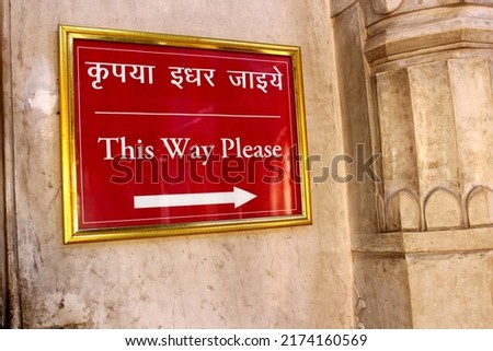 Red sign board hanging on the wall. Sign says this way please. Sign board in hindi and english showing direction to move. Arrow showing to move in left direction.